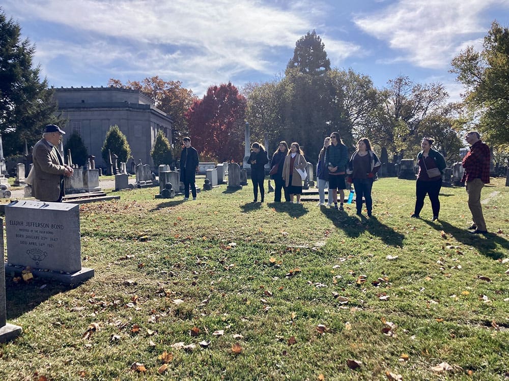 Students stand at a distance from Elijah Bond's tombstone